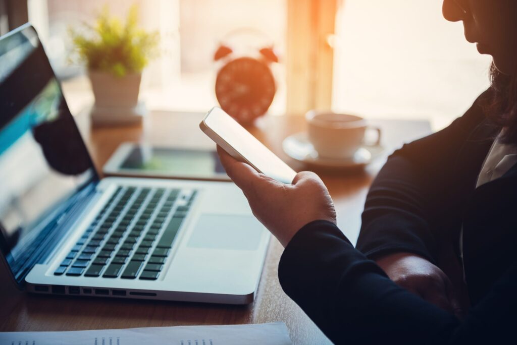 A person sitting at a desk using a smartphone, with a laptop, a cup of coffee, an alarm clock, and a potted plant nearby. Sunlight is shining in through a window.