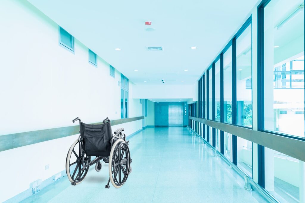 An empty wheelchair sits in the middle of a bright, clean hospital hallway with large windows and a view outside.