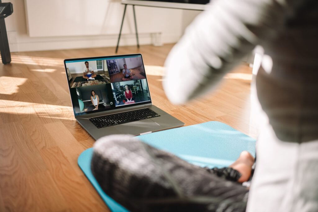 A person sits on a yoga mat in front of a laptop displaying a virtual yoga class with four participants on screen, all practicing yoga together in different home settings.