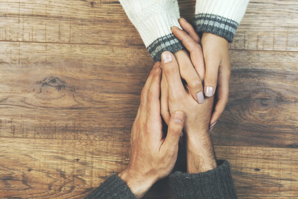 Two people sitting at a wooden table gently hold hands, one persons hands resting comfortingly on top of the others, conveying support and care. Both are wearing long-sleeved sweaters.