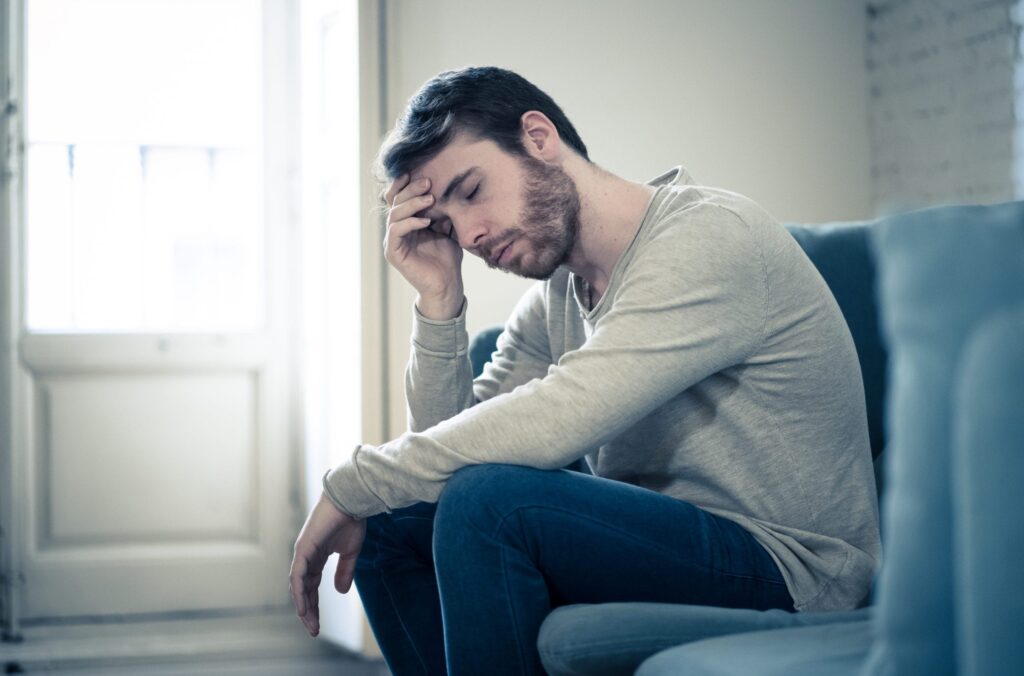 A young man sits on a couch indoors, leaning forward with his elbow on his knee and his hand on his forehead, appearing stressed or deep in thought.