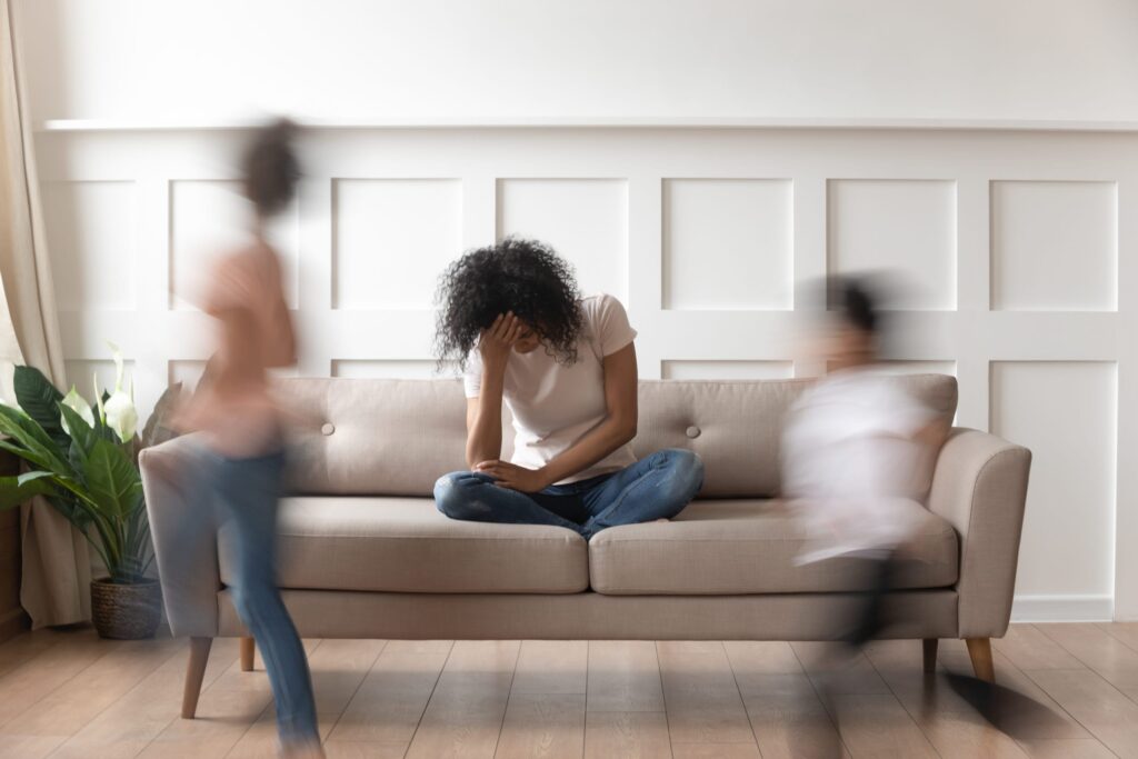 A woman sits on a sofa with her head in her hand, looking stressed, while two children run past her, creating a blurred motion effect. The room is bright with a plant in the corner.