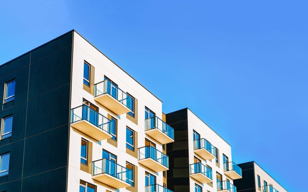 Modern apartment building with multiple glass balconies, white and dark exterior walls, set against a clear blue sky.