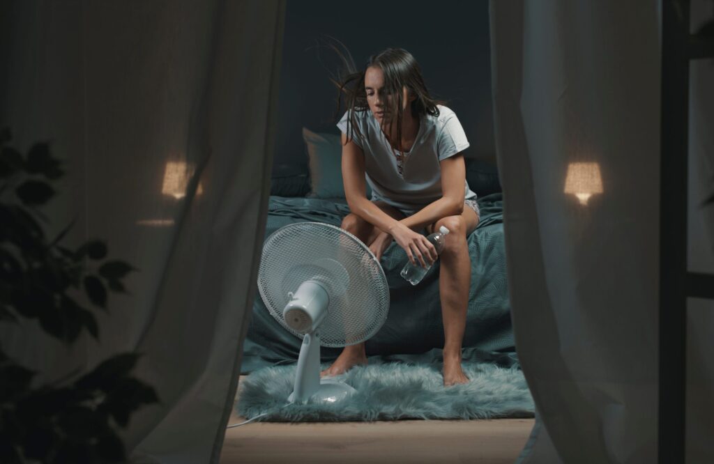 A woman sits on the edge of a bed at night, holding a water bottle and facing an electric fan, with her hair being blown by the fan. The room is dimly lit and framed by curtains.