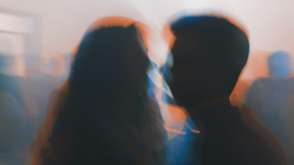Blurry image of two people facing each other closely, possibly speaking or about to kiss, with a colorful background and soft lighting creating a dreamy, abstract effect.