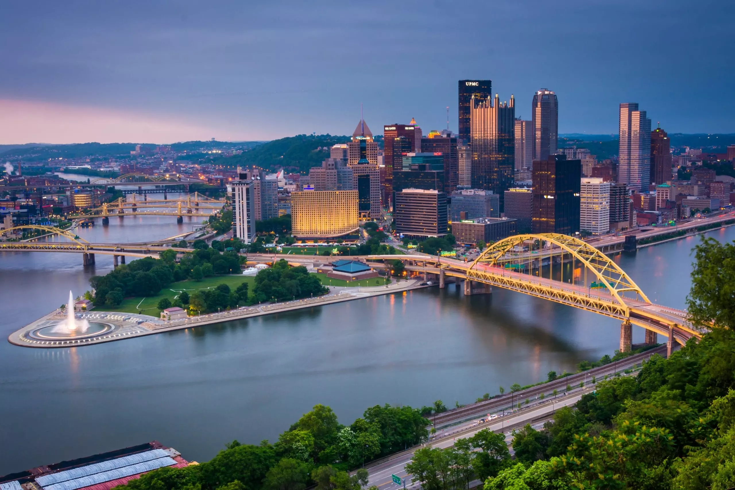 A view of downtown Pittsburgh at dusk shows tall buildings, yellow bridges crossing rivers, Point State Park with its fountain, and lush green trees in the foreground.