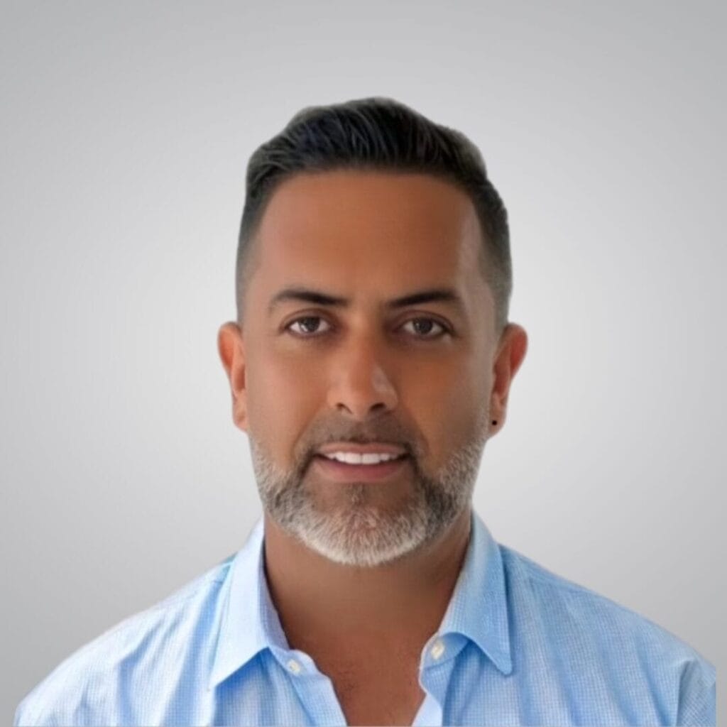 A man with short dark hair and a trimmed salt-and-pepper beard, wearing a light blue collared shirt, smiles slightly in front of a plain light gray background.