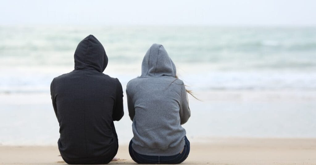 Two people, sitting solemnly by the beach, worried about their daughter, wondering "what is enabling in addiction?" and what some signs of enabling parents actually are.