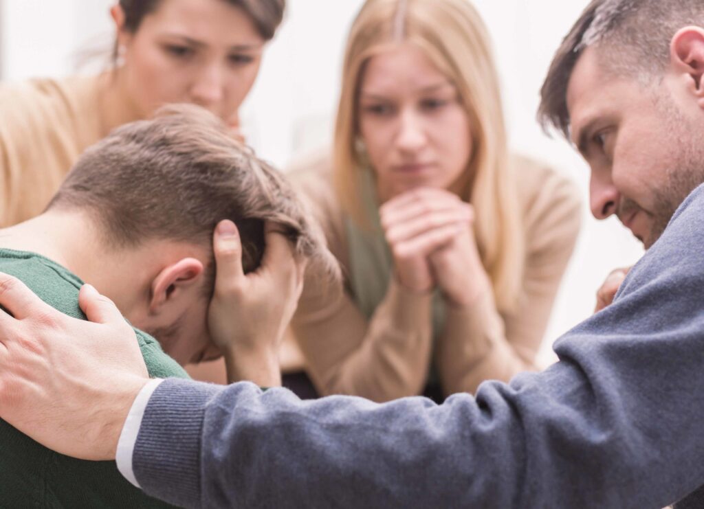 Four people sit close together. One person appears distressed, with head in hands, while the others surround him, showing concern and support. The scene conveys empathy and comfort in a difficult moment.