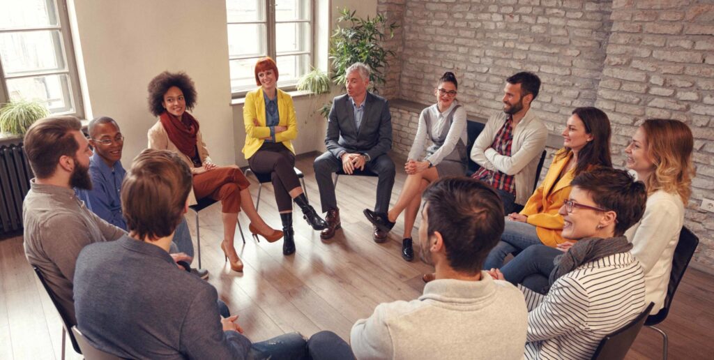 A diverse group of people sit in a circle in a bright room, engaged in conversation, suggesting a team meeting or support group session. Everyone appears attentive and relaxed.