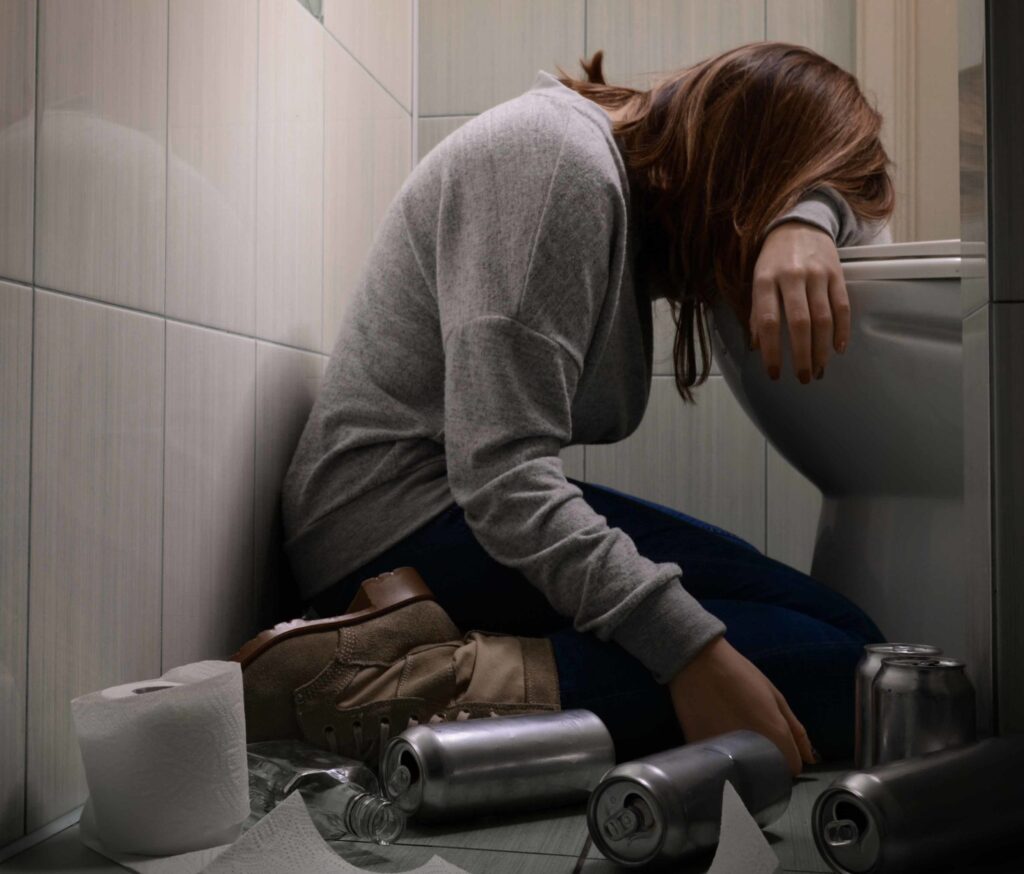 A woman sits on the bathroom floor, slumped over a toilet with her head resting on her arm. Empty cans and a roll of toilet paper are scattered on the floor around her.