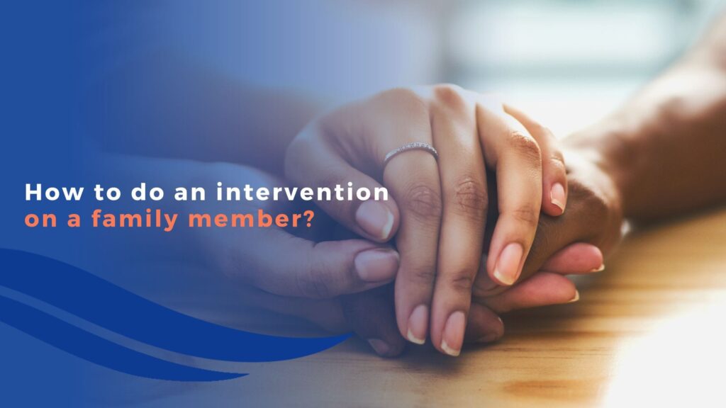 Two hands gently holding each other on a wooden table, with text overlay: How to do a family intervention? Blue wave design on the left side of the image.