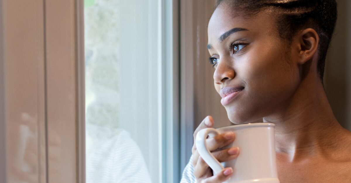 woman in recovery, staring in peaceful contemplation out a window with mug in hand.