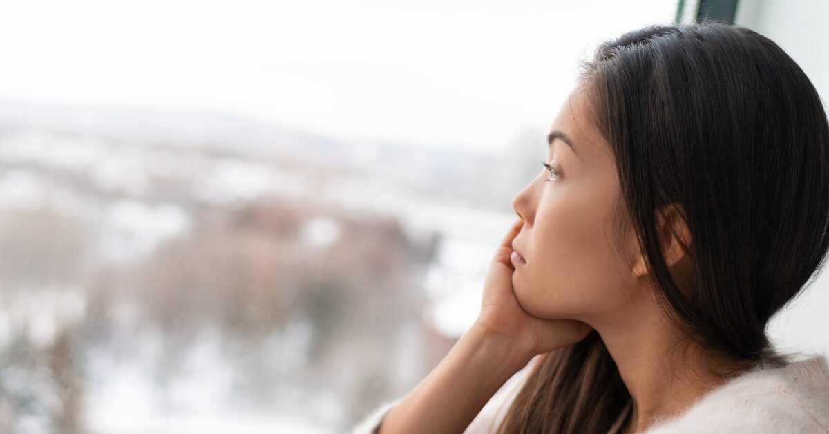 Woman suffering from SAD and addiction looks glumly out a window at winter scenery.