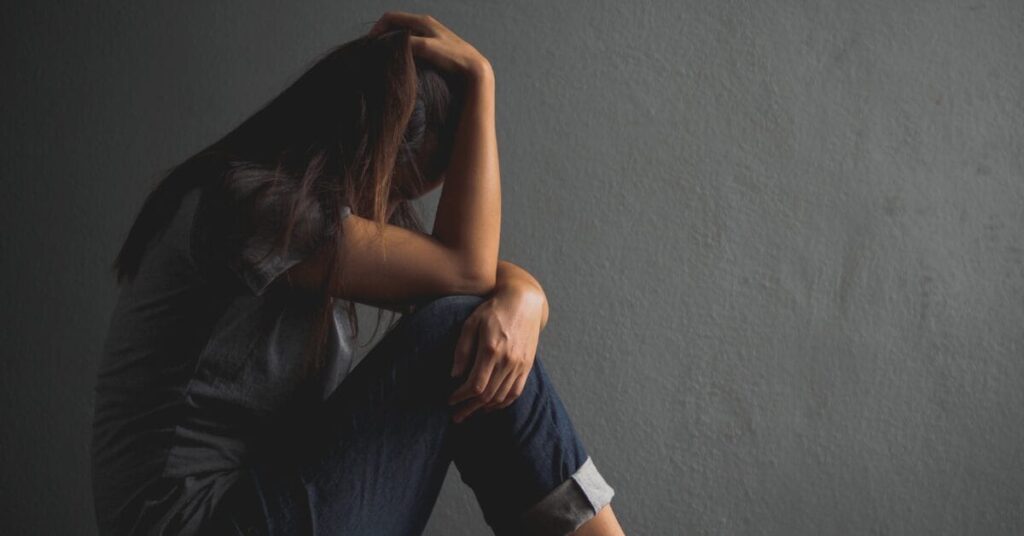A woman crouches in an empty gray room, turning her face away from the camera, representing the importance of alcohol awareness month.