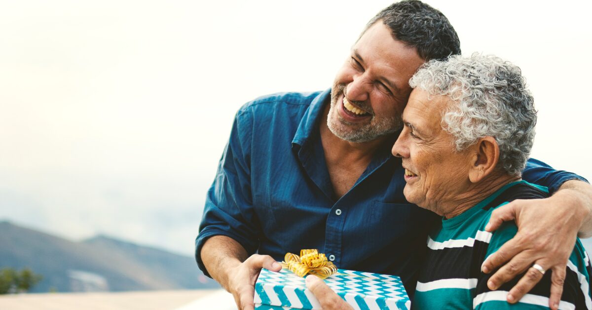 A middle-aged son hands his father a chevron-printed present while embracing him, with a mountain scene in the background. Men's sobriety leads to stronger relationships and increased health.
