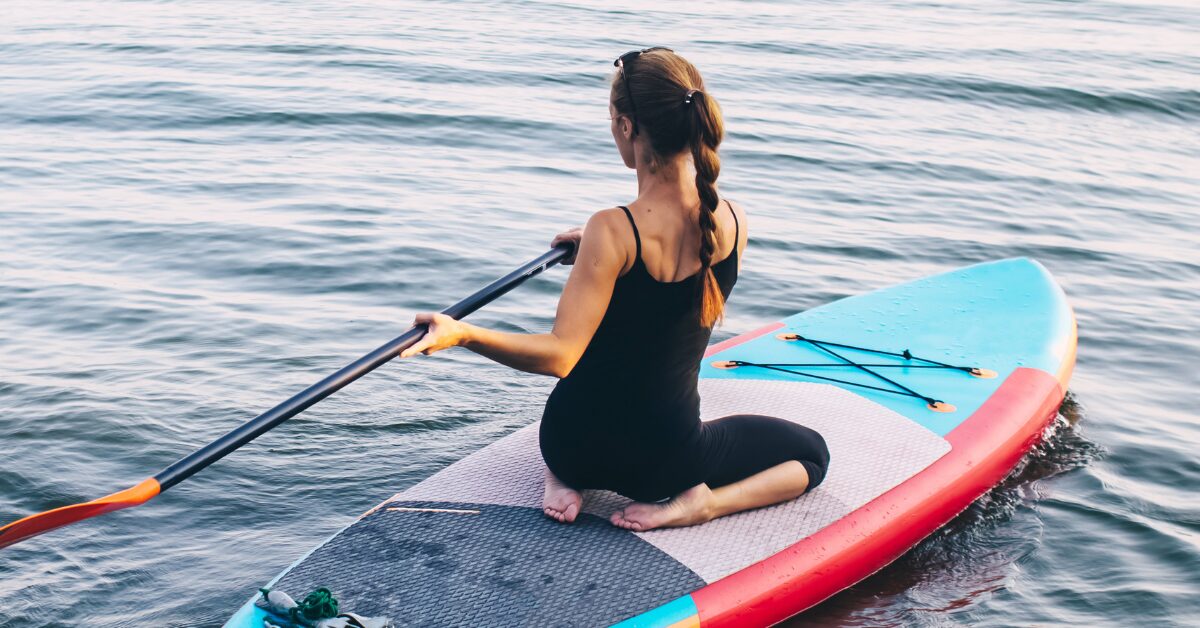 A woman in a black singlet with her back to camera sits on a standup paddleboard in the middle of a rippling body of water, engaging in adventure therapy.