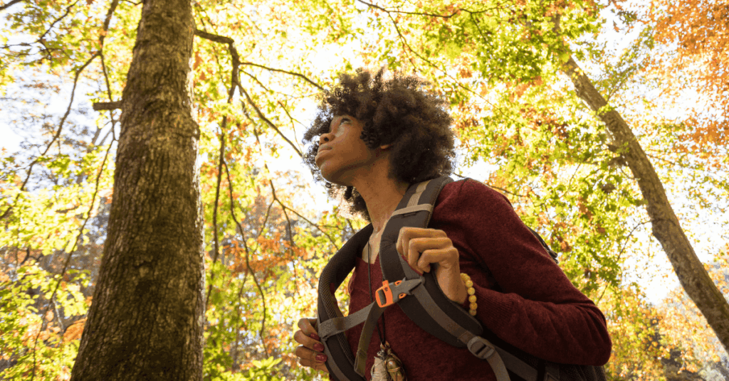 A woman holds onto her backpack straps as she hikes through the woods during the fall. Physical activity is one of many important coping strategies to reduce Fall stress and relapse risk.