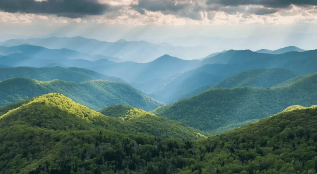 Sunlight streams through clouds over rolling, tree-covered mountains, creating soft rays and layers of blue and green ridges that stretch into the distance under a dramatic sky.
