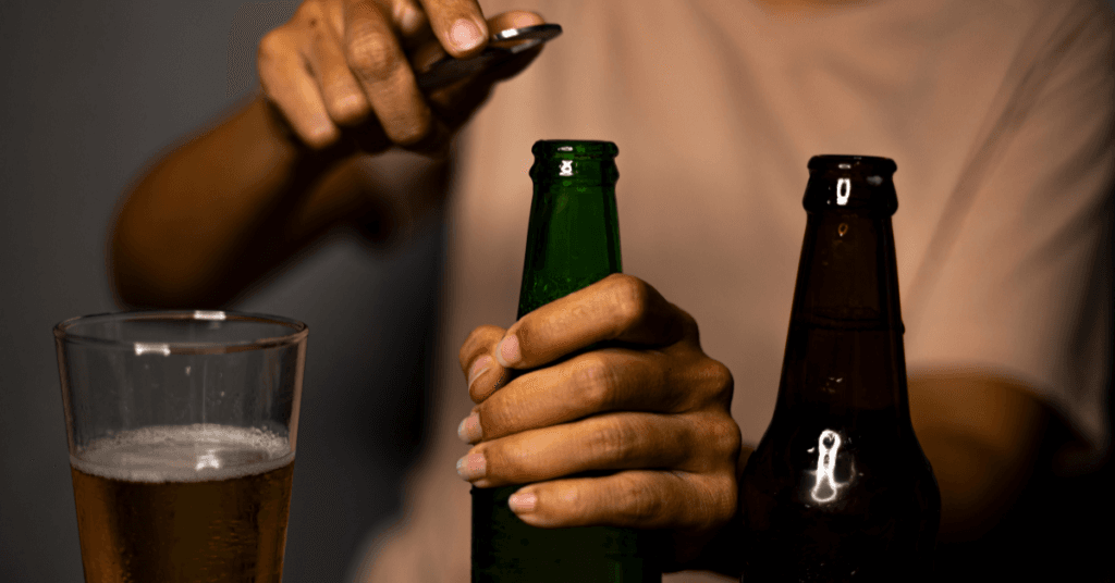 A man with alcohol use disorder opening a green beer bottle, with multiple other drinks around him.
